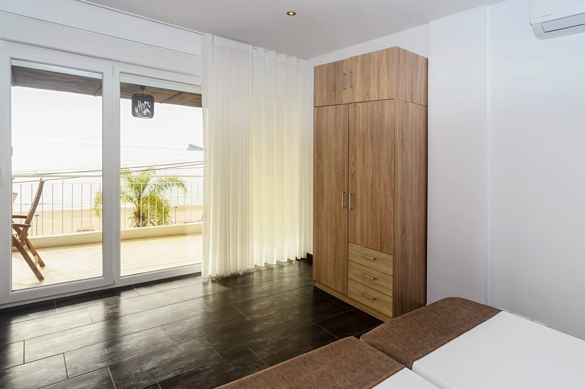 A room with a sliding glass door leading to a balcony, a wooden wardrobe, and a brown sofa.