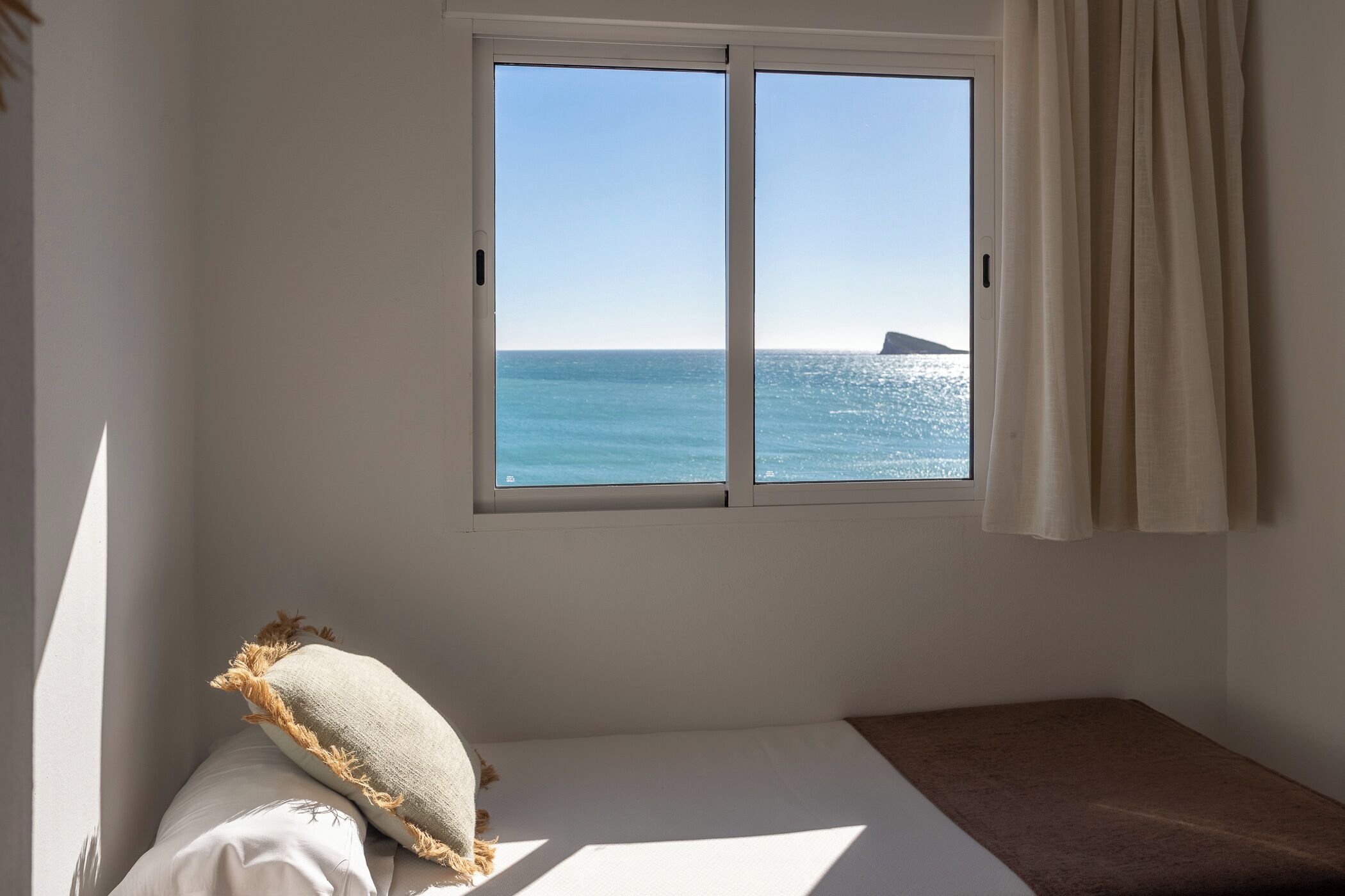 A room with a bed, a window with a view of the sea, and a curtain.