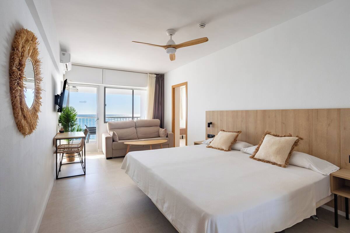 Ein Hotelzimmer mit Bett, Deckenventilator, Fernseher, Balkon mit Aussicht und Essbereich mit Tisch und Stühlen.