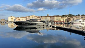En småbåtshamn med flera yachter förtöjda, en stilla vattenyta som speglar båtarna och byggnaderna, och en klar himmel med enstaka moln.