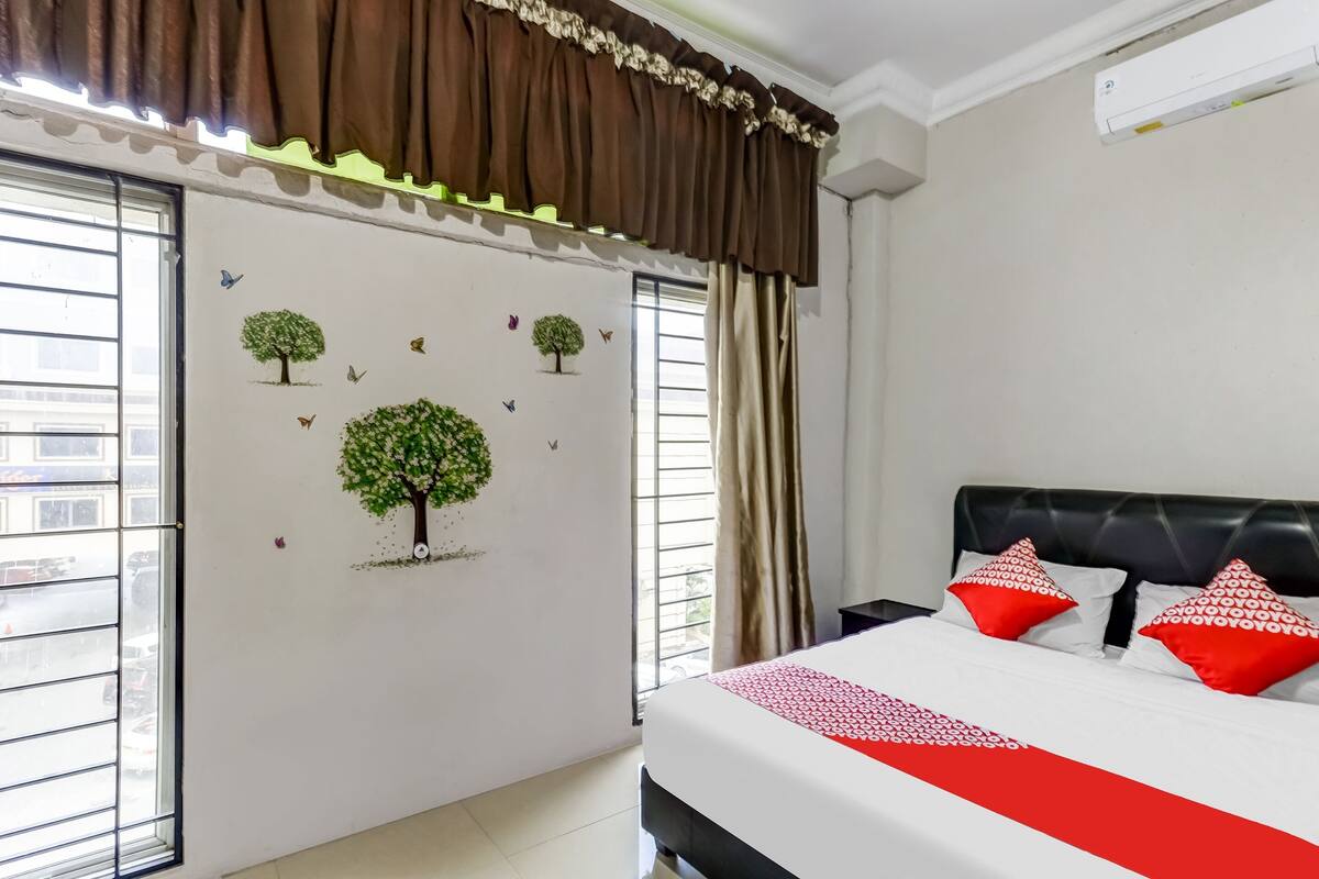 Ein Schlafzimmer mit einem Bett, roten und weißen Kissen, einem Fenster mit Jalousien sowie Wanddekorationen mit Bäumen und Schmetterlingen.