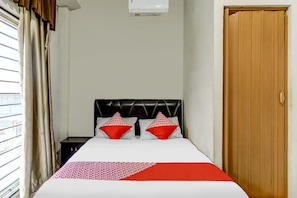 Hotelkamer met een bed, twee rode kussens, een raam met gordijnen, een houten deur en een wandairconditioner.