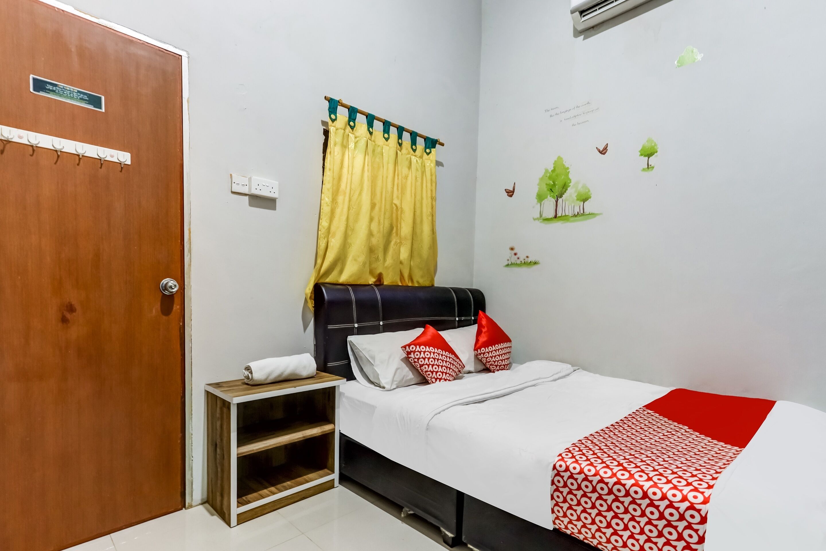 A bedroom with a bed, a nightstand, a wooden shelf, a door, and a wall with a window and wall stickers.