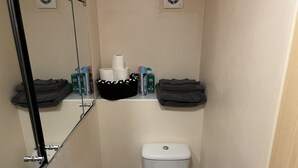 A small bathroom with a toilet, a sink, and a mirror. There are toiletries and towels on a shelf above the toilet. - Flat In Willen - Milton Keynes suitable for contractors and family staycations  (Milton Keynes)