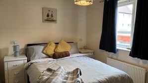 A bedroom with a bed, nightstands, a lamp, a painting, and a window with curtains. - Flat In Willen - Milton Keynes suitable for contractors and family staycations  (Milton Keynes)
