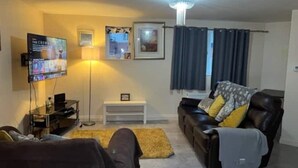 A living room with a flat-screen TV, a standing lamp, a sofa with cushions, a small white side table, and a ceiling light fixture. - Flat In Willen - Milton Keynes suitable for contractors and family staycations  (Milton Keynes)