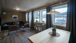 Una habitación acogedora con paredes y techo de madera, un sofá negro, una mesa de comedor de madera con sillas y un ventanal con vistas a un estacionamiento y montañas.