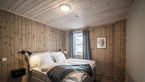 Un dormitorio con techo de madera, una cama con ropa blanca, un plaid gris, una lámpara de noche y una ventana con cortinas.