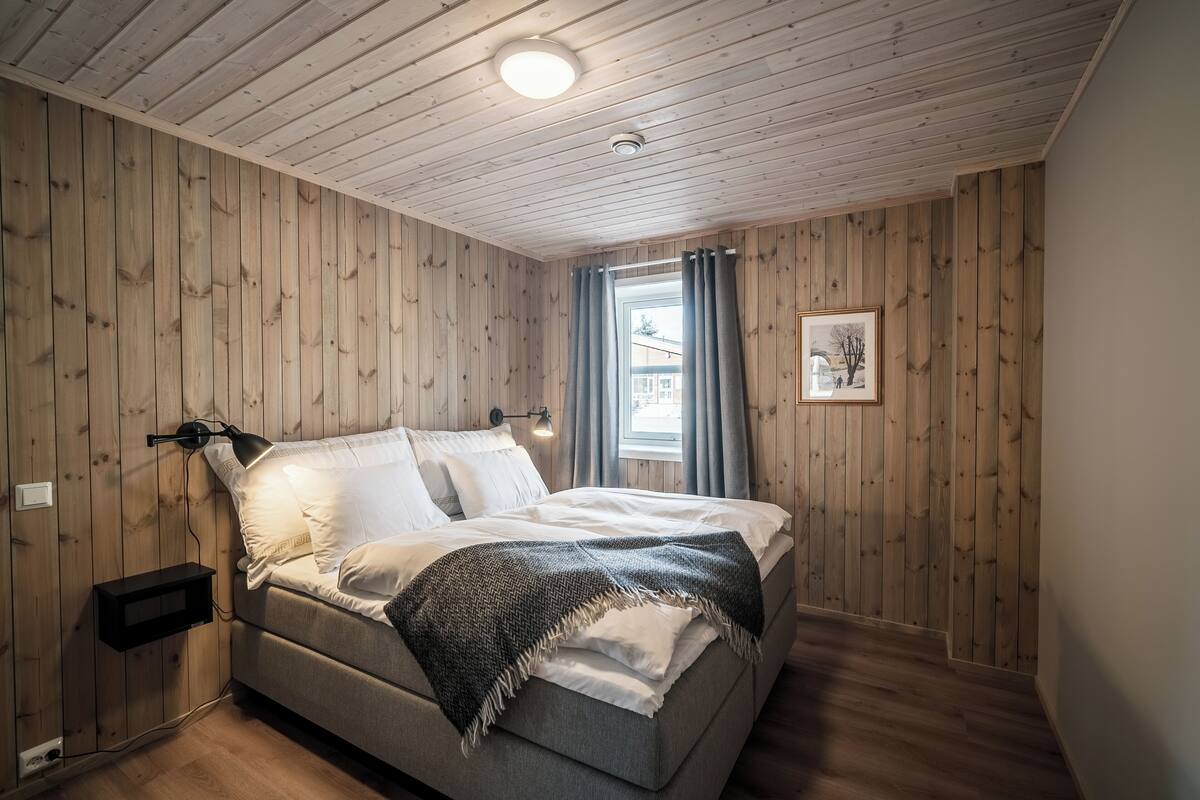 A bedroom with a wooden ceiling, a bed with white linens, a gray throw blanket, a bedside lamp, and a window with curtains.