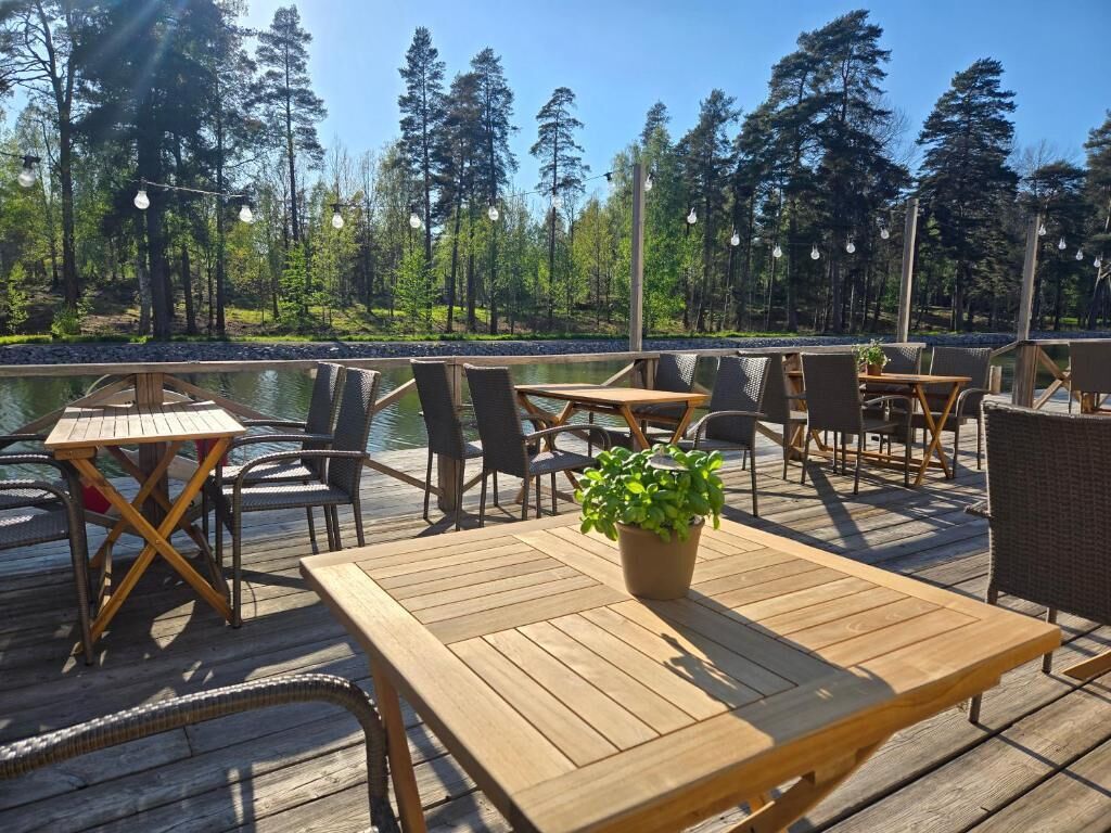 A wooden deck with tables and chairs, a small body of water, and a forested background.