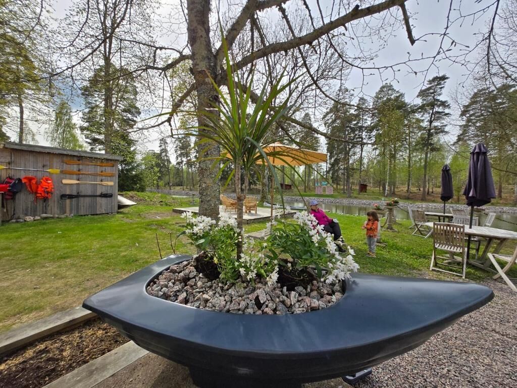 Ein Gartenbereich mit einem zentralen Pflanzgefäß, Gartenmöbeln und einer Holzkonstruktion im Hintergrund.