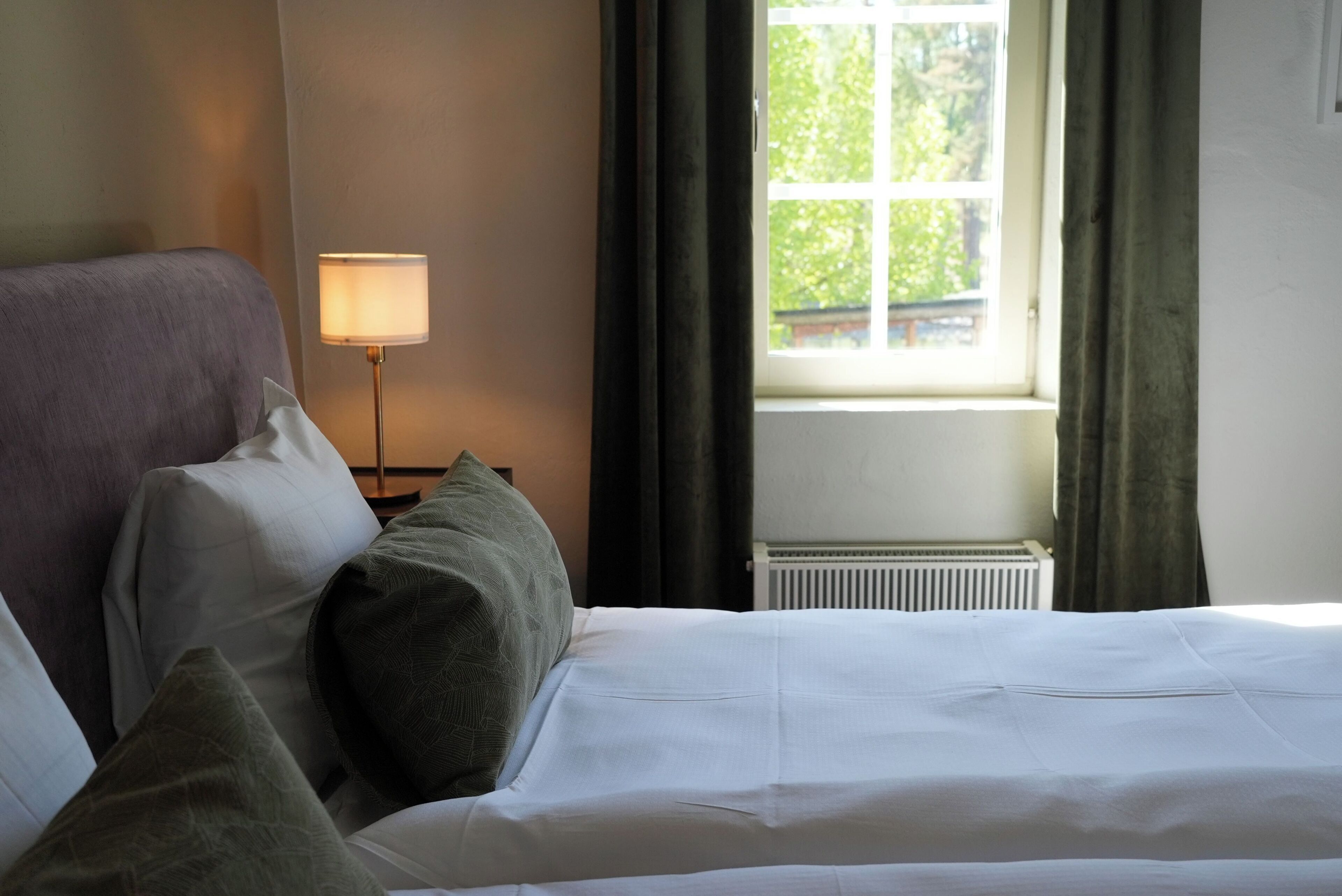 A neatly made bed with white linens, a bedside lamp, and a window with curtains.