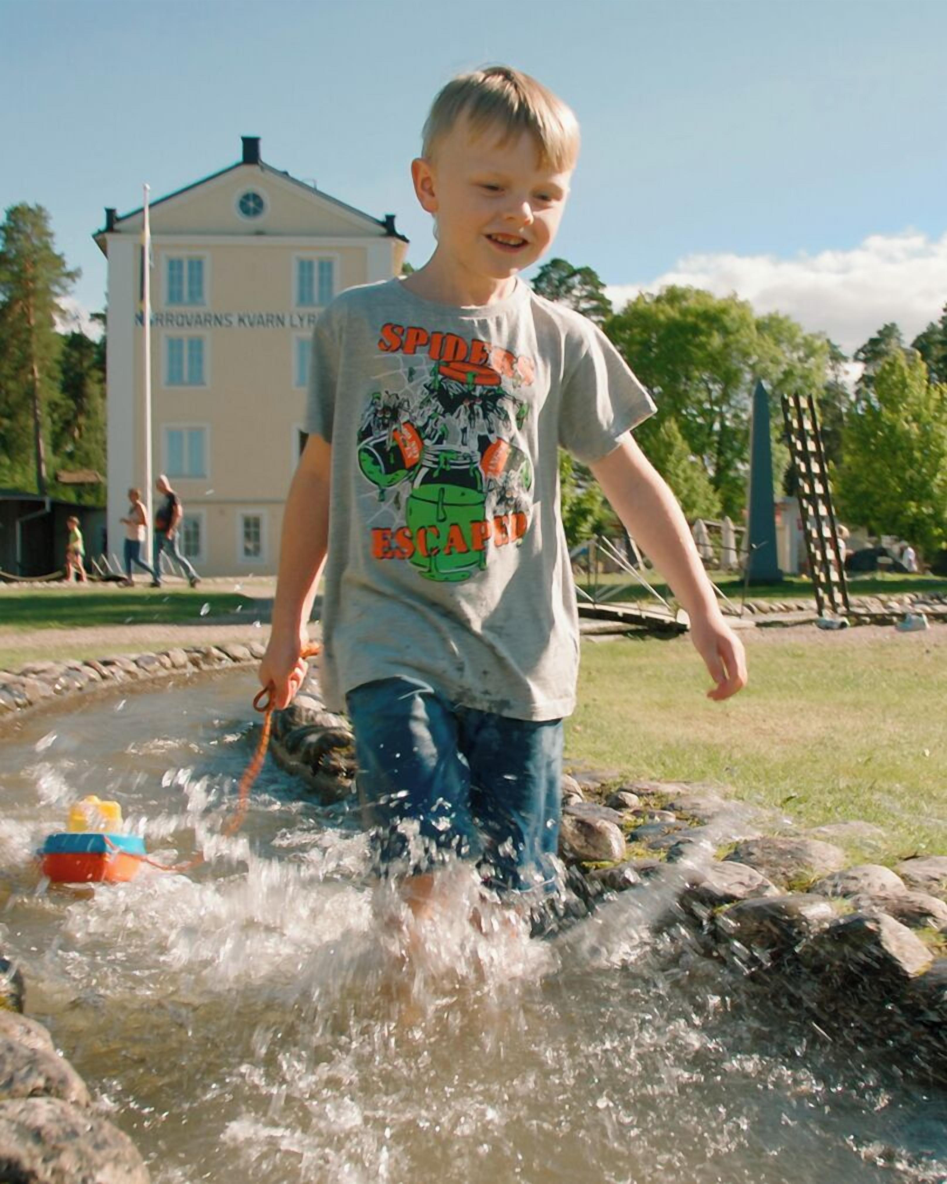 Un enfant joue dans une fontaine ou un autre jeu d’eau, avec un bâtiment en arrière-plan.