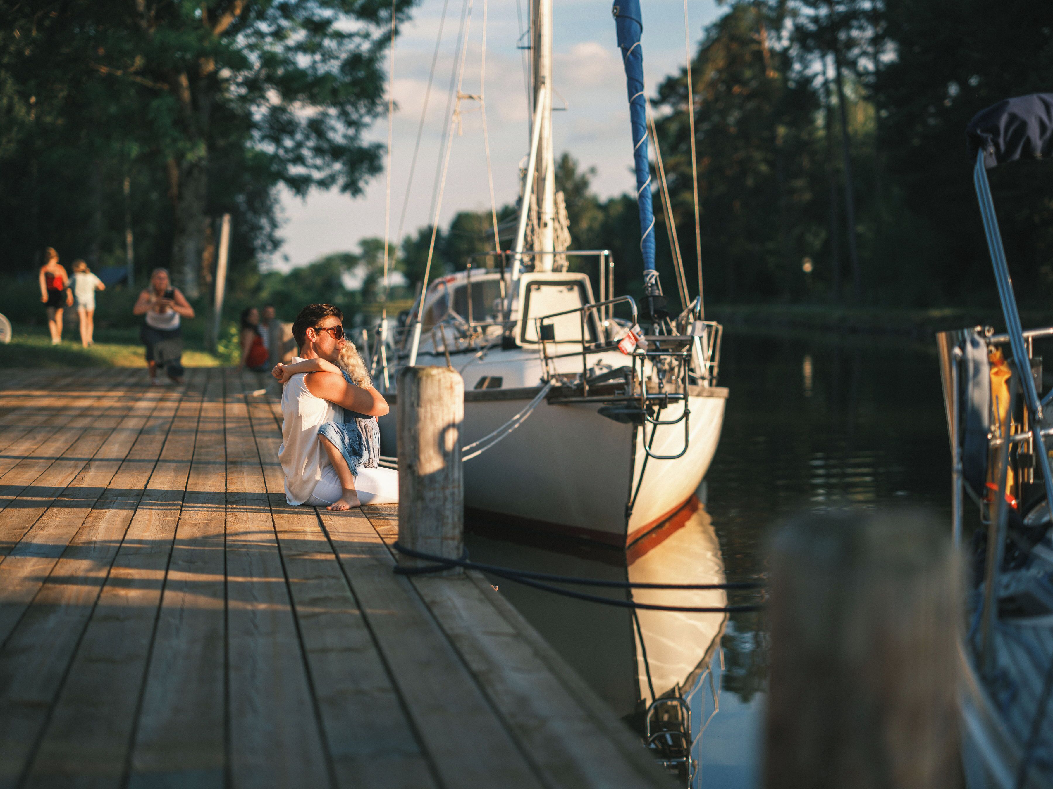 A wooden dock with a sailboat moored at a pier, surrounded by calm water and trees.