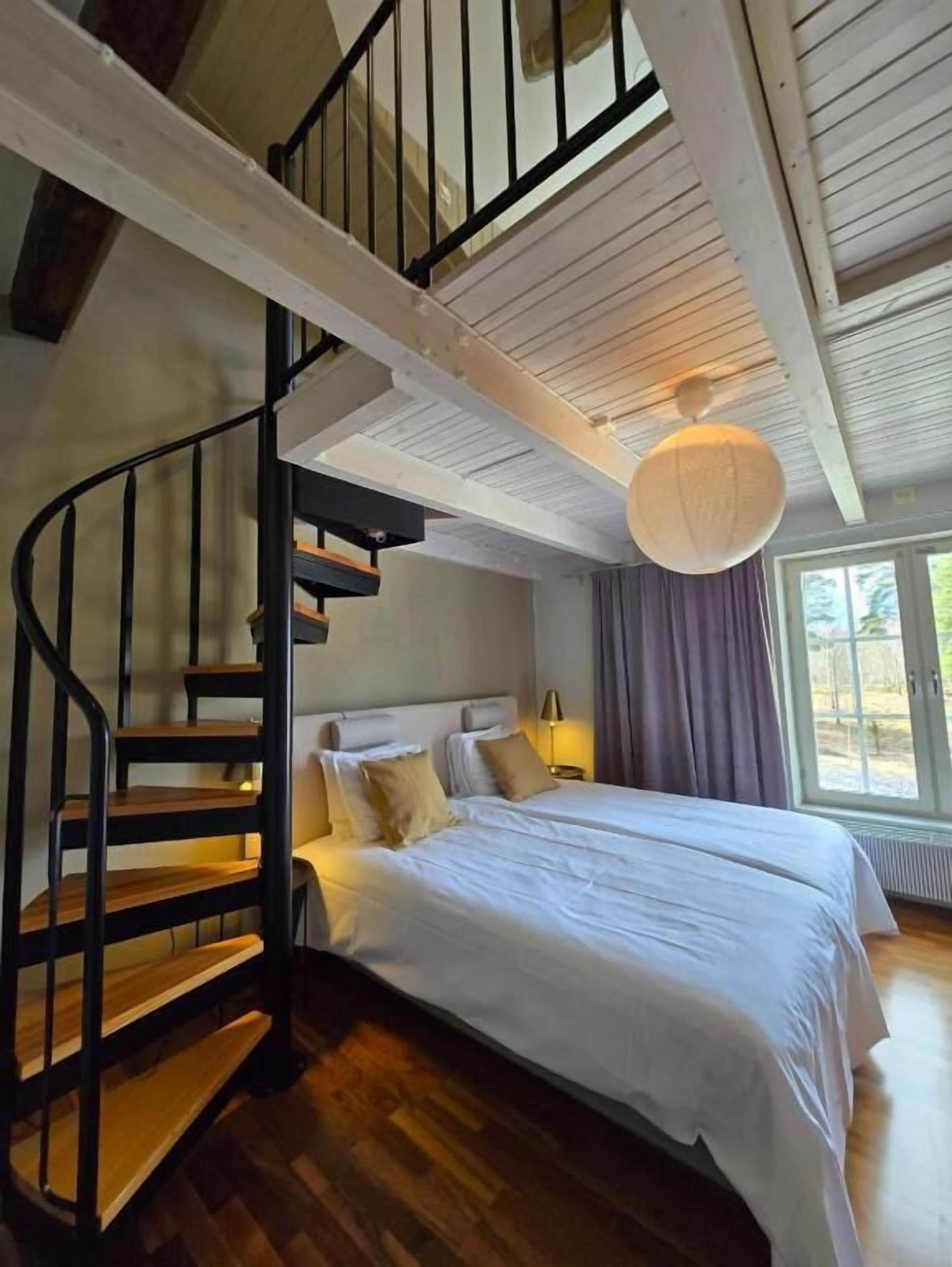 A bedroom with a wooden staircase, a bed with white linens, and a large window.