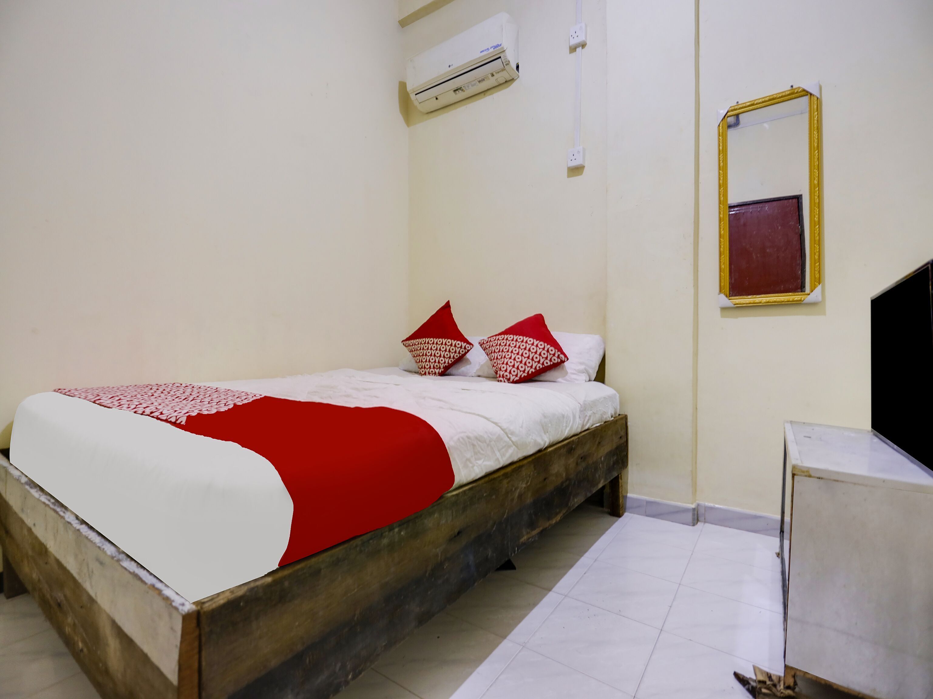 Ein Schlafzimmer mit einem hölzernen Bett, weißer Bettwäsche, roten und weißen Kissen, einer Klimaanlage, einem Spiegel und einem kleinen Tisch.