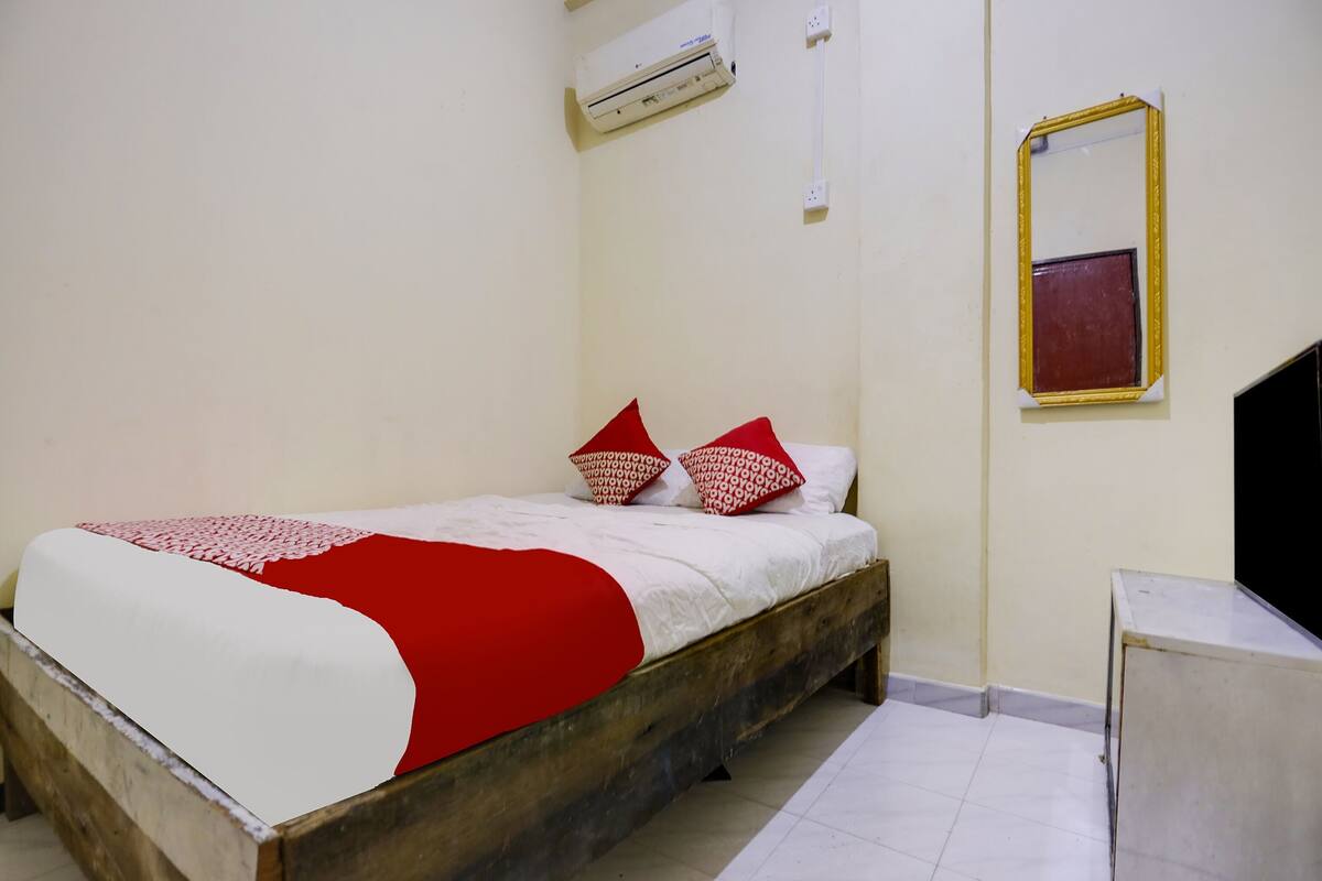 A bedroom with a wooden bed, white bedding, red and white pillows, an air conditioning unit, a mirror, and a small table.