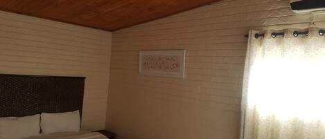 A bedroom with a wooden ceiling, a bed with white bedding, a headboard, a wall-mounted air conditioning unit, and a window with a curtain.
