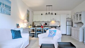 A modern living room with a white sofa, a white armchair, and a coffee table. The room has a kitchen in the background with white cabinets and a refrigerator. - House by the sea, 2 km from Baratti gulf & Etruscan archaeological park (Stazione di Populonia)