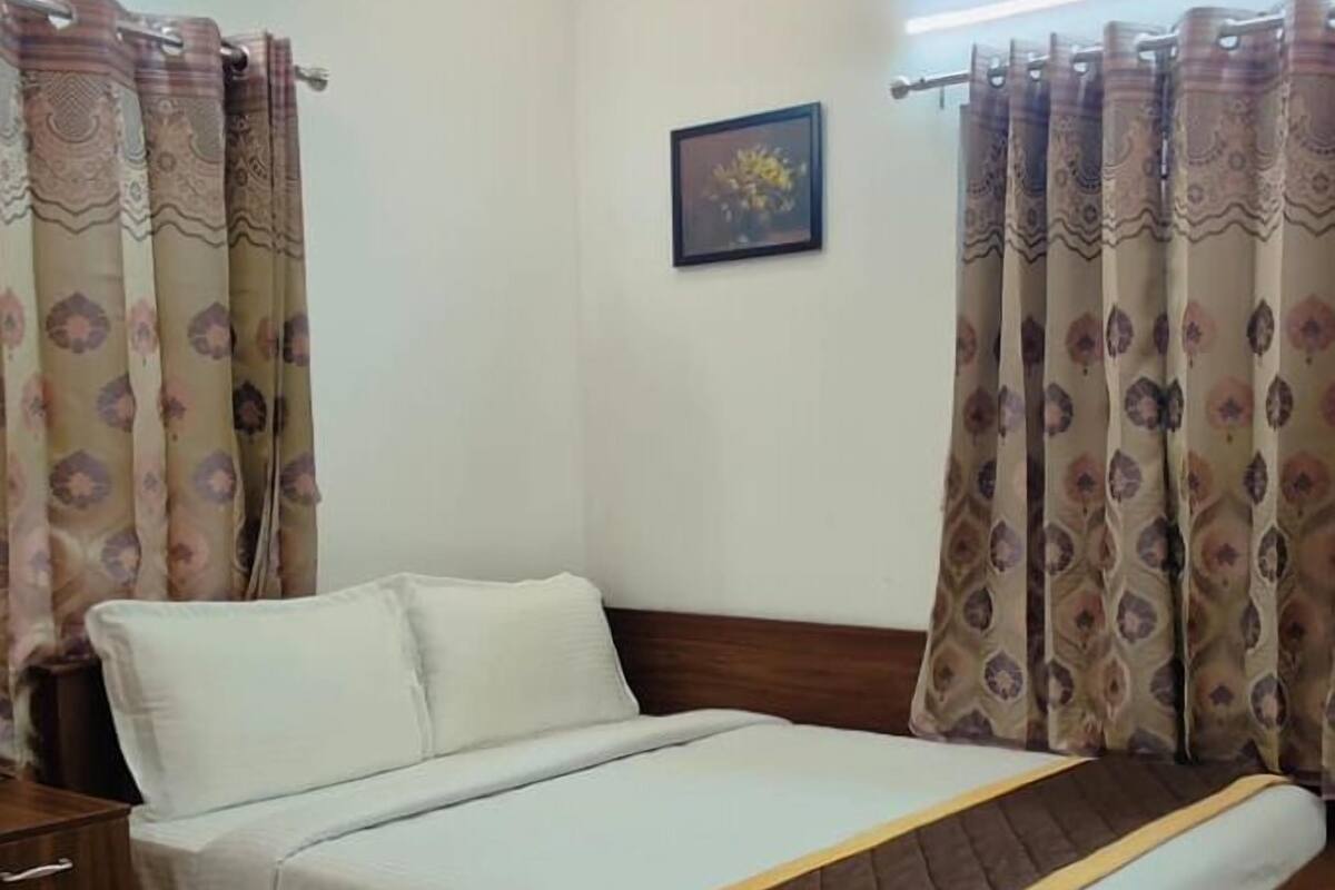 A neatly made bed with a wooden frame and a white and brown bedspread. There is a nightstand with a lamp on it to the left of the bed. The room has a tiled floor and a picture hanging on the wall above the bed.