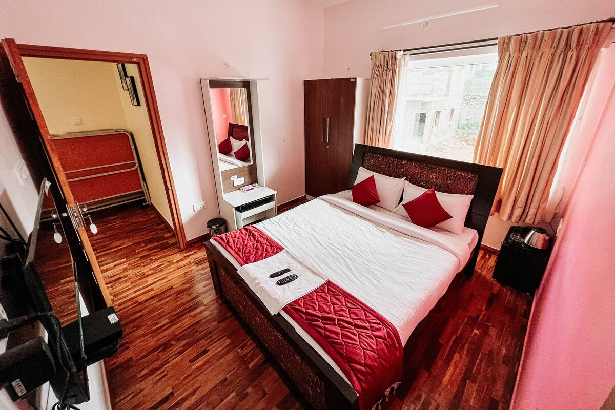 A hotel room with a wooden floor, a bed with red and white bedding, a wardrobe, a mirror, and a window with curtains.