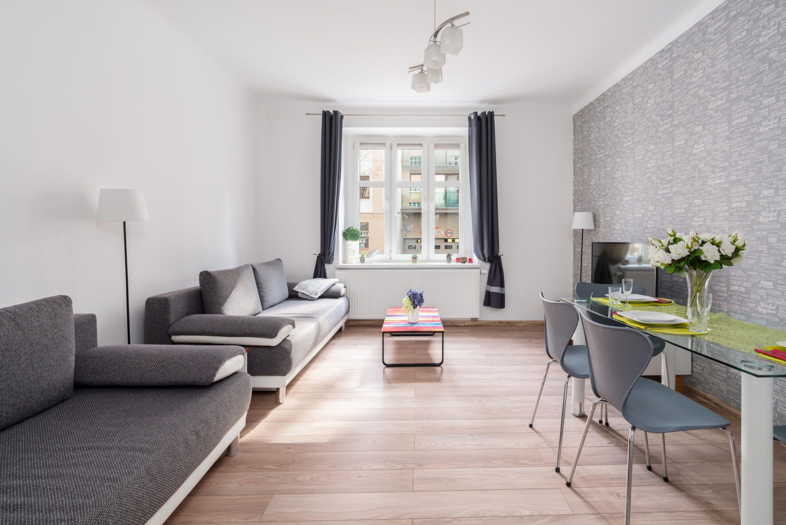 A bright living room with a gray sofa, a matching armchair, a coffee table, and a dining table with chairs. The room has a large window that allows plenty of natural light.