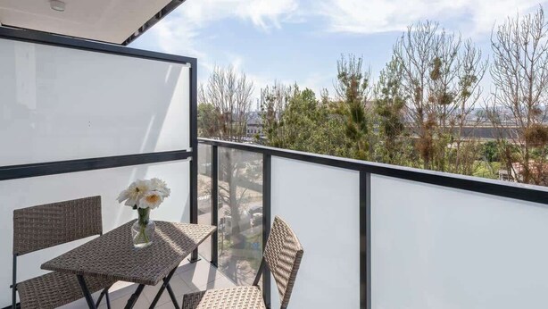 Un balcon agrémenté de meubles en osier et d’un vase de fleurs, donnant sur un quartier résidentiel avec des arbres et des maisons.