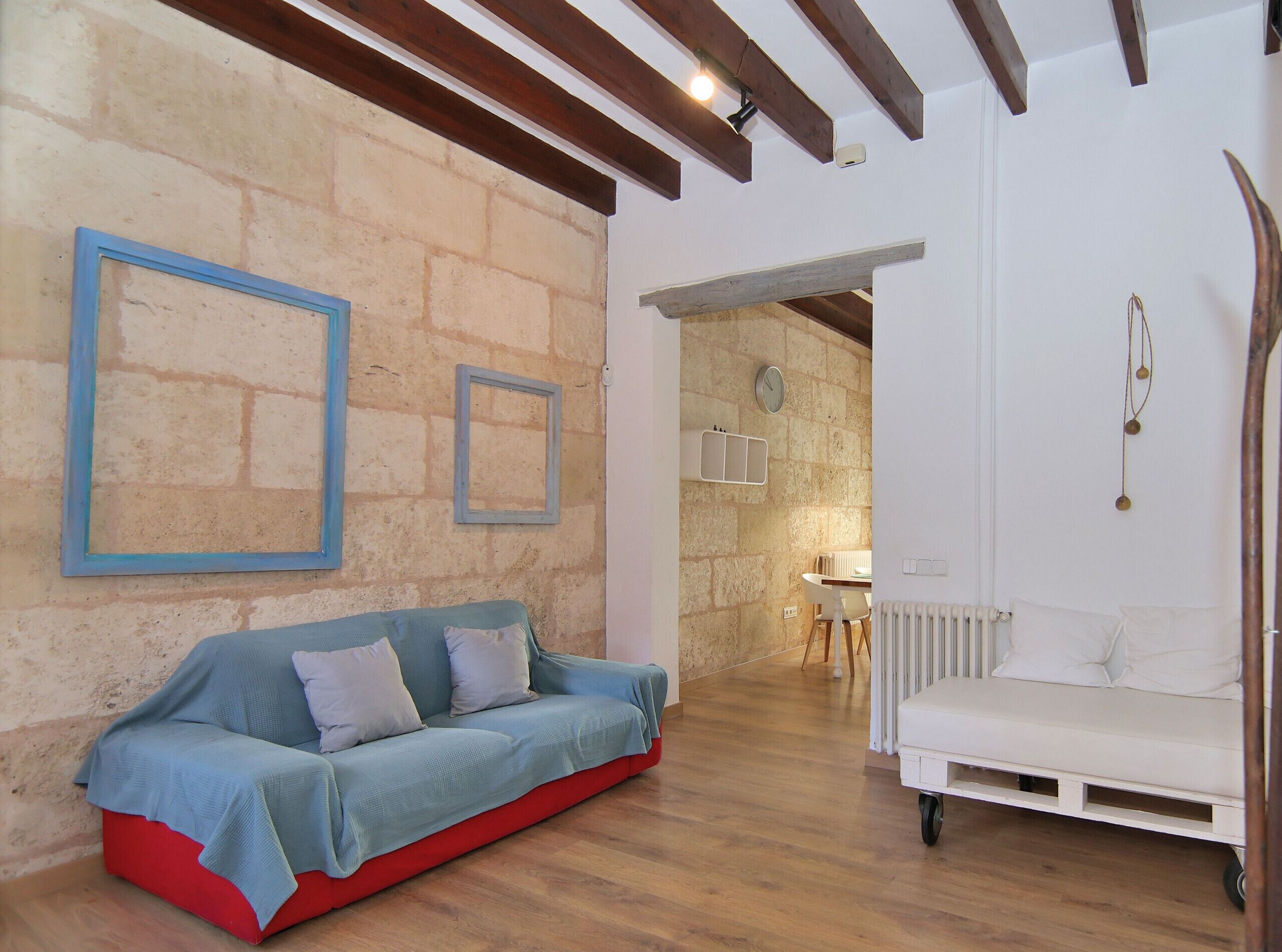 A cozy living room with a blue sofa, a white sofa, and a red ottoman. There are two framed pictures on the wall, a clock, and a string of beads.