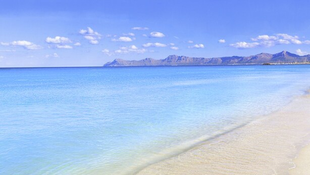 Une plage à l’eau d’un bleu cristallin, avec une chaîne de montagnes en arrière-plan.