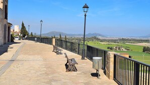 Un sentier pavé de pierres, agrémenté de bancs et d’une rambarde en métal, offrant une vue sur des champs verdoyants et des montagnes lointaines.