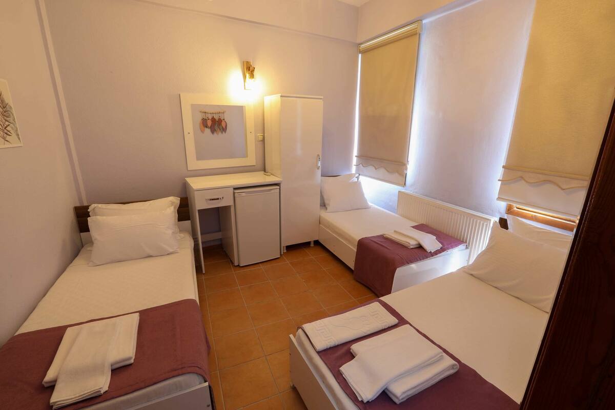 Una habitación pequeña y limpia con dos camas individuales, un escritorio, un refrigerador pequeño y una ventana con cortinas.