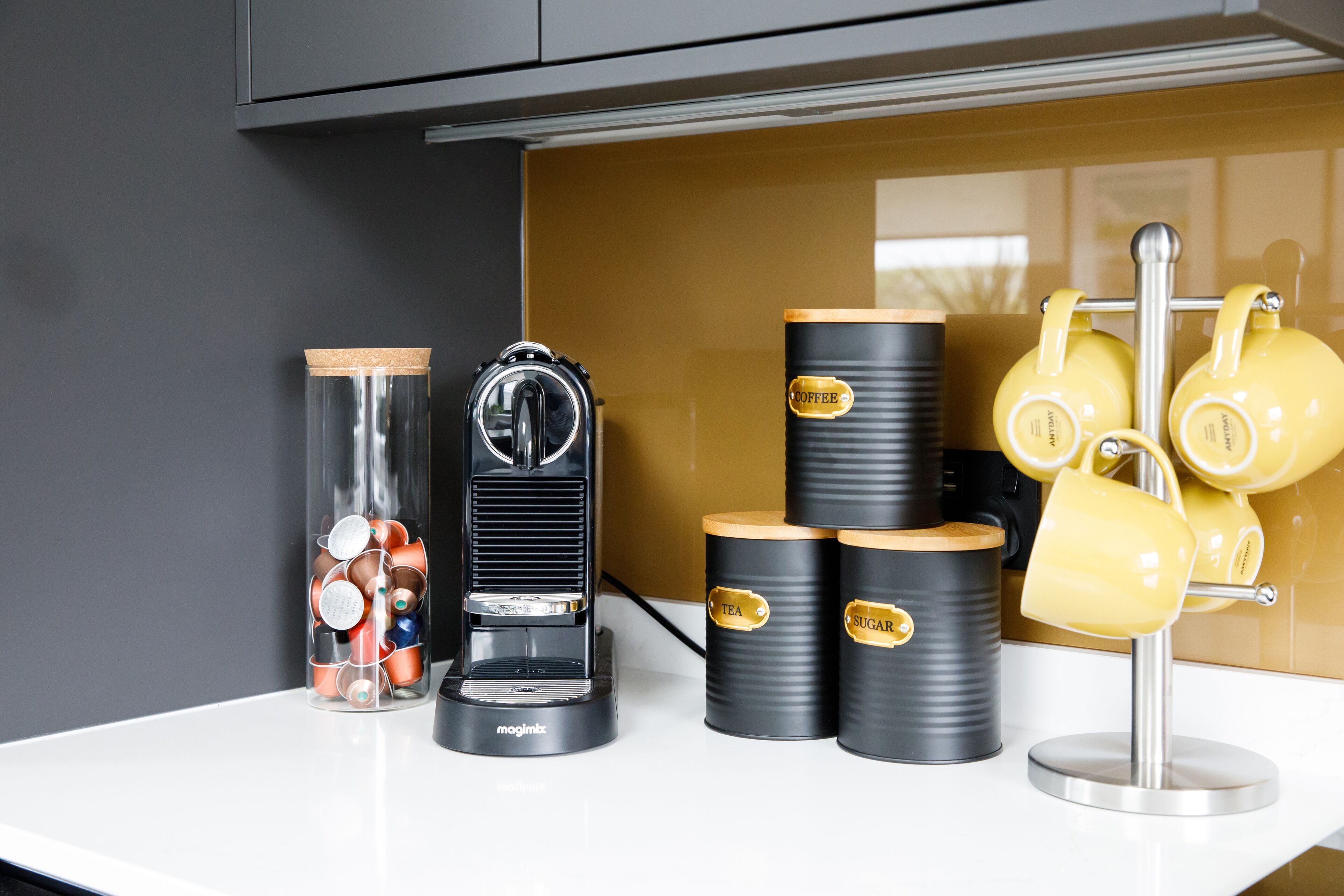 A kitchen counter with a coffee maker, a glass jar with lids, and two containers labeled 'tea' and 'sugar'.