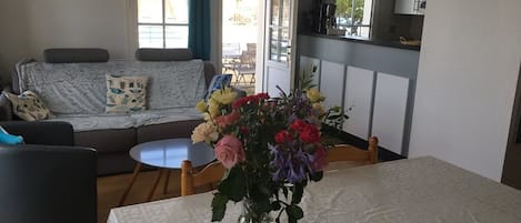 A living room with a sofa, a dining table with a vase of flowers, and a kitchen area in the background.