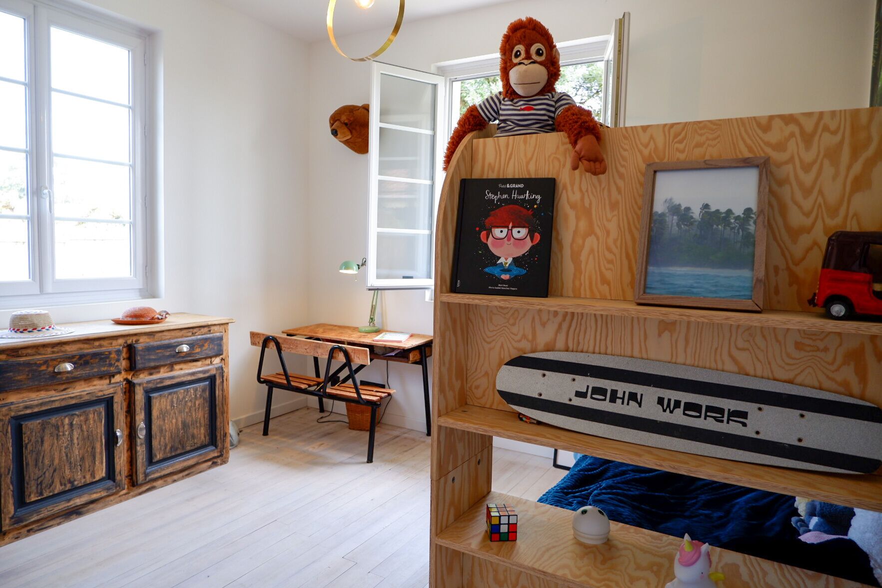 Une chambre d’enfant avec un bureau, une chaise et un singe en peluche. On y trouve une bibliothèque avec un cadre photo et une voiture jouet.