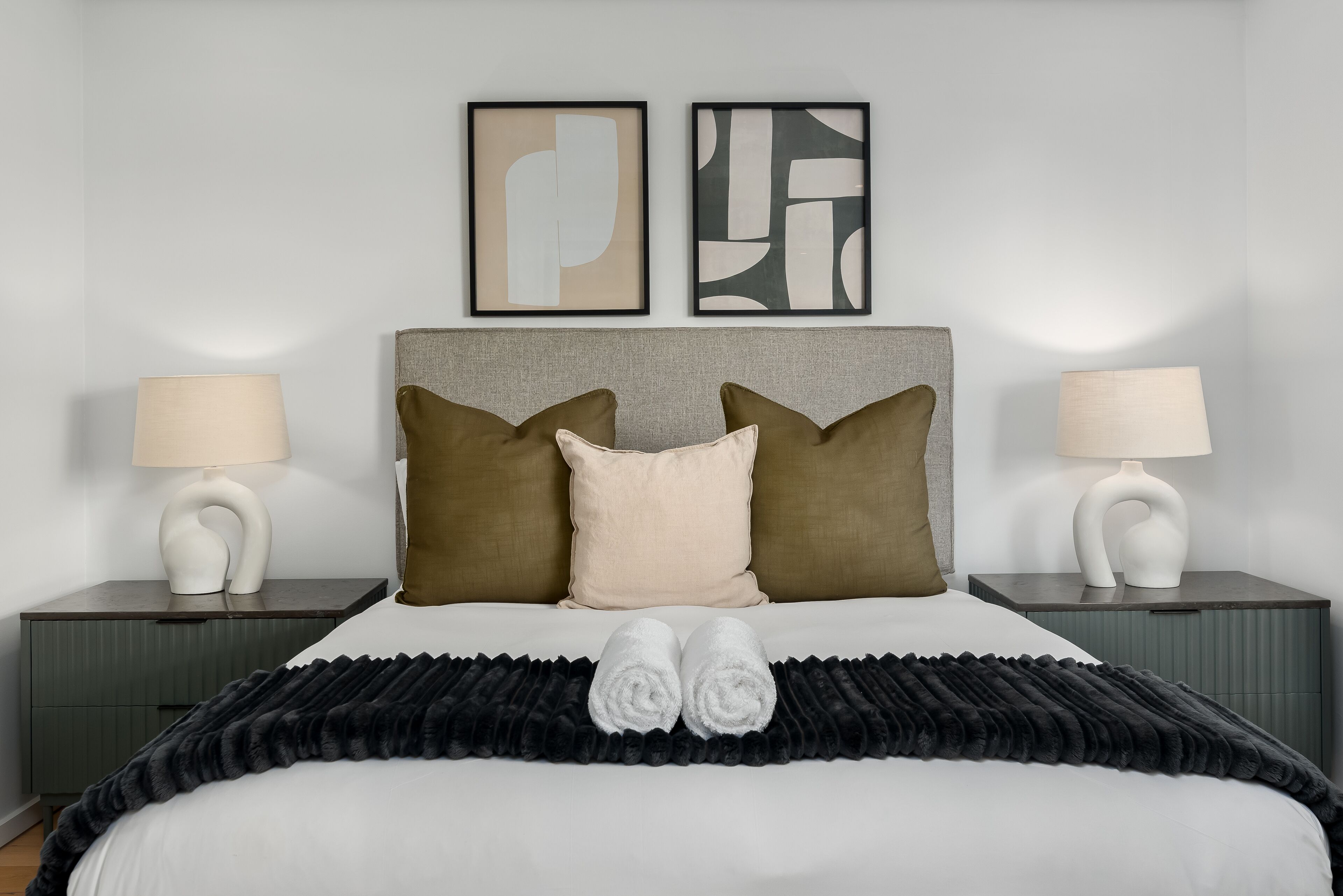 A neatly made bed with two pillows, a black throw blanket, and two rolled towels. There are two bedside tables with lamps and framed artwork above the bed.
