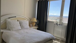 Habitación de hotel con una cama grande, una mesita de noche con lámpara, una ventana con cortinas y vistas a edificios y montañas.