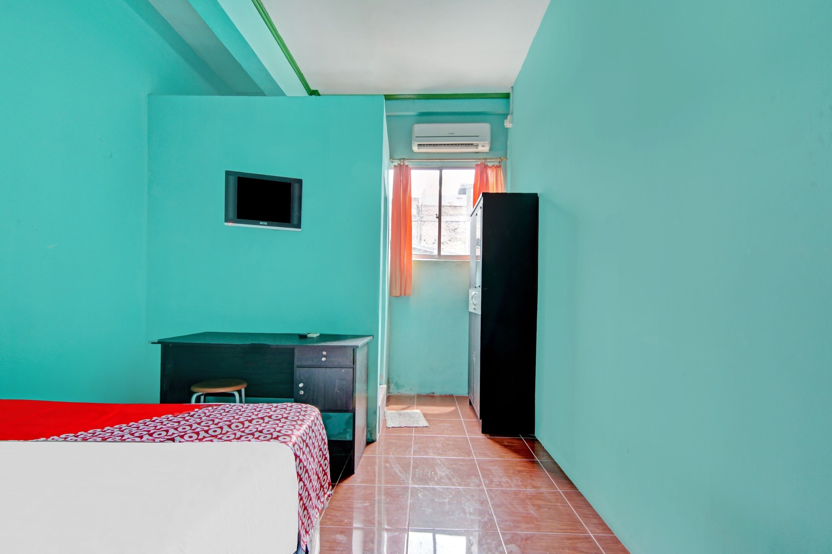 A room with teal walls, a bed with a red and white patterned bedspread, a desk with a chair, a black wardrobe, and an air conditioning unit near a window with orange curtains.