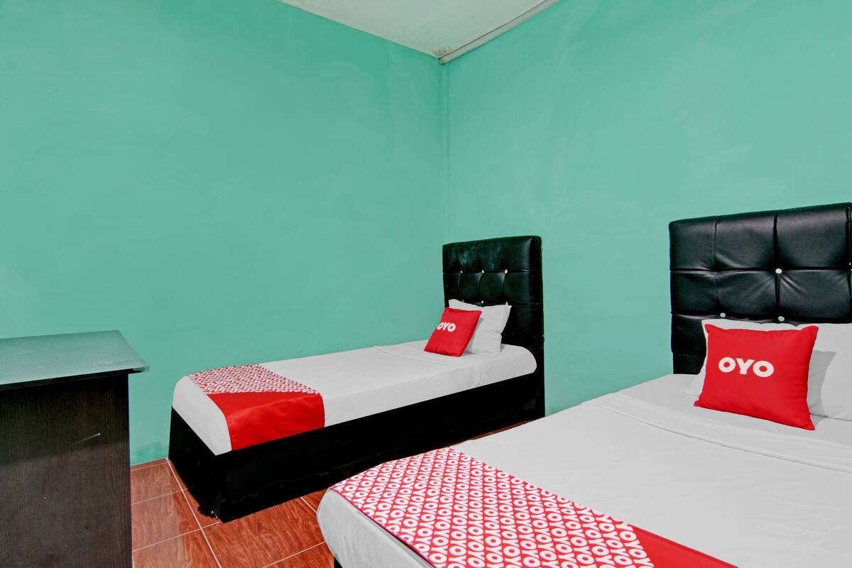 A hotel room with two beds, each with a black headboard and red pillows. The room has a teal wall and a wooden nightstand.