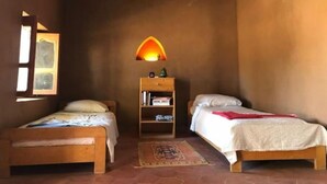Una habitación pequeña y acogedora con dos camas de madera, una mesita de noche con libros, una lámpara de pared y una ventana con vistas.