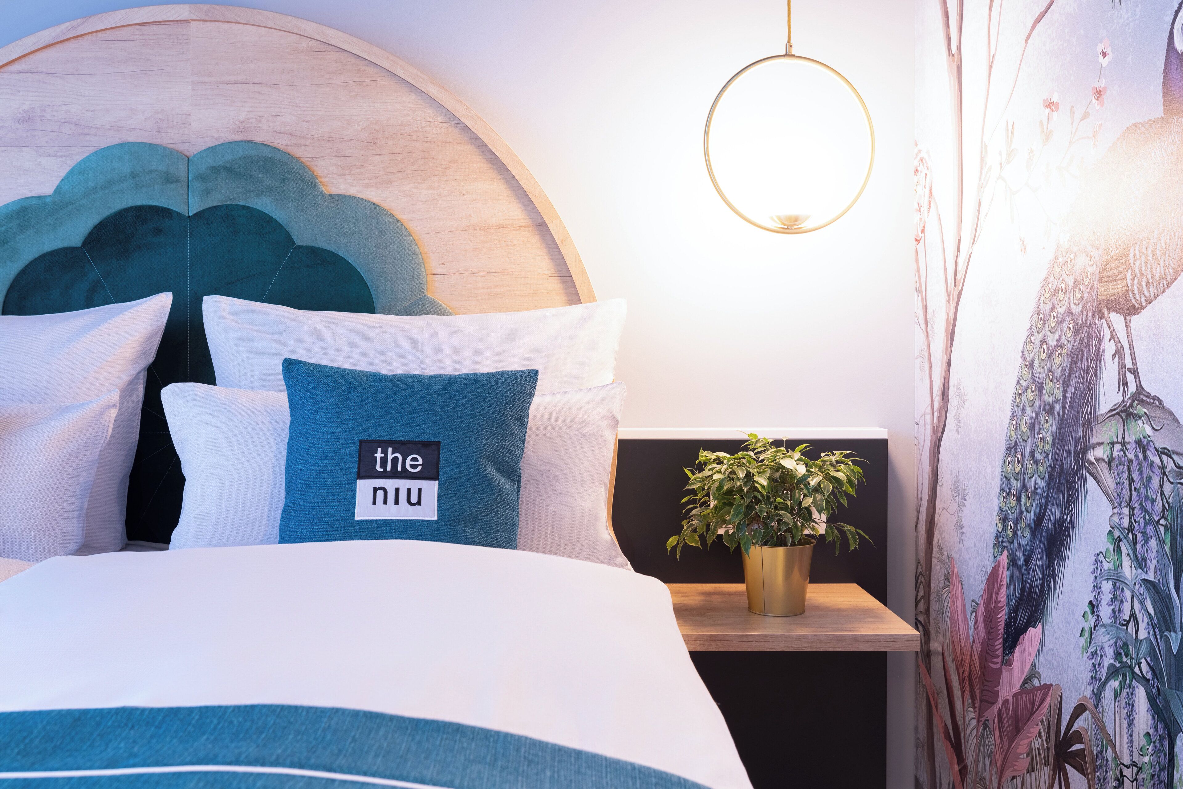 Ein ordentlich bezogenes Bett mit Kopfteil, ein Nachttisch mit Pflanze und eine Wand mit einem Wandbild zum Thema Natur.