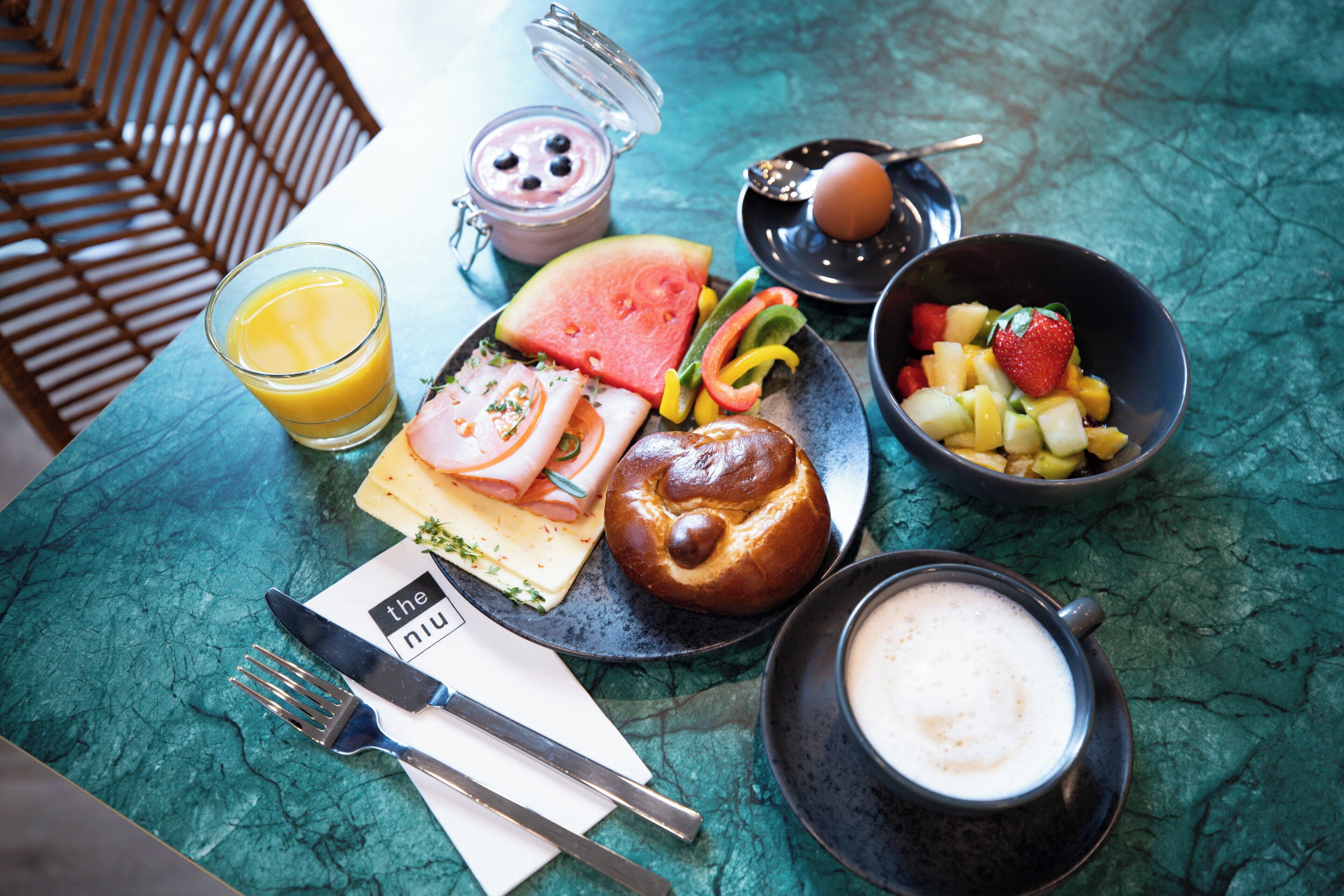 Un desayuno con un vaso de zumo de naranja, un bol de fruta, un pastel, una taza de café y un tarro pequeño de yogur.