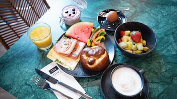 Un desayuno con un vaso de zumo de naranja, un bol de fruta, un pastel, una taza de café y un tarro pequeño de yogur.