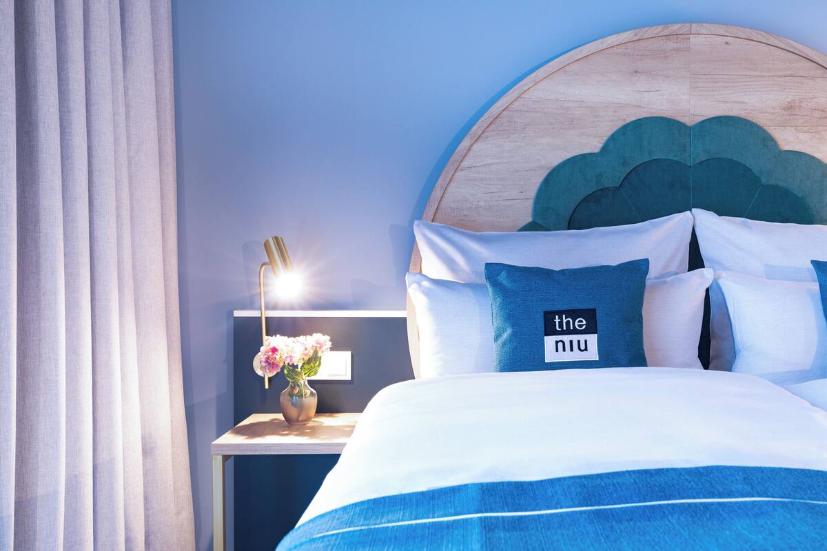 Una cama bien hecha con una colcha azul y blanca a rayas, un cabecero de madera y una mesita de noche con un florero y una lámpara.