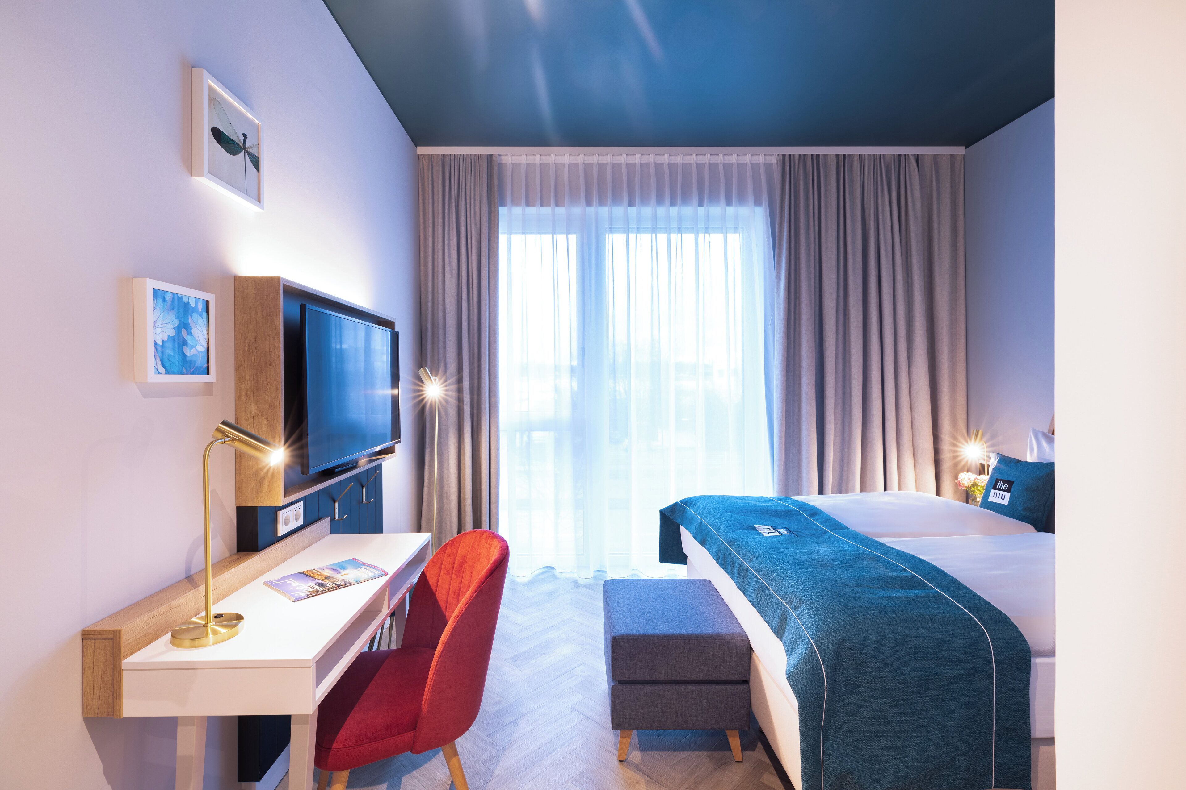 A modern hotel room with a desk, chair, television, and a bed with a blue bedspread.