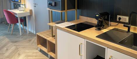 Una cocina moderna con estufa integrada, cafetera y pileta. Se observa un escritorio con silla y un televisor al fondo.