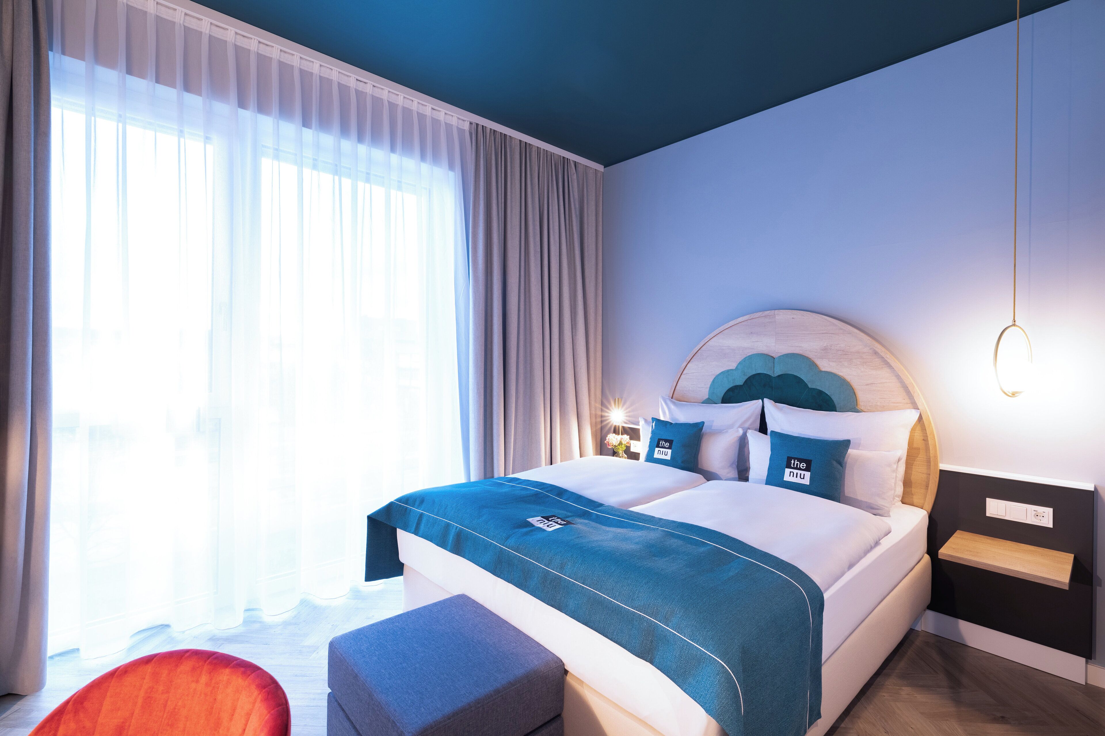 A modern hotel room with a large bed, a bedside table with a lamp, and a view of the outdoors through sheer curtains.