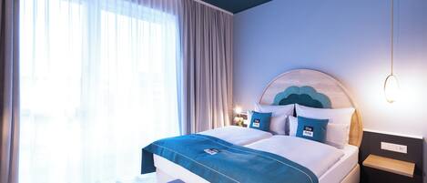 A modern hotel room with a large bed, a bedside table with a lamp, and a view of the outdoors through sheer curtains.