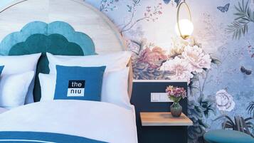 Een slaapkamer met een bed, kussens, een nachtkastje, een vaas met bloemen, behang met bloemen en een hangende lamp.