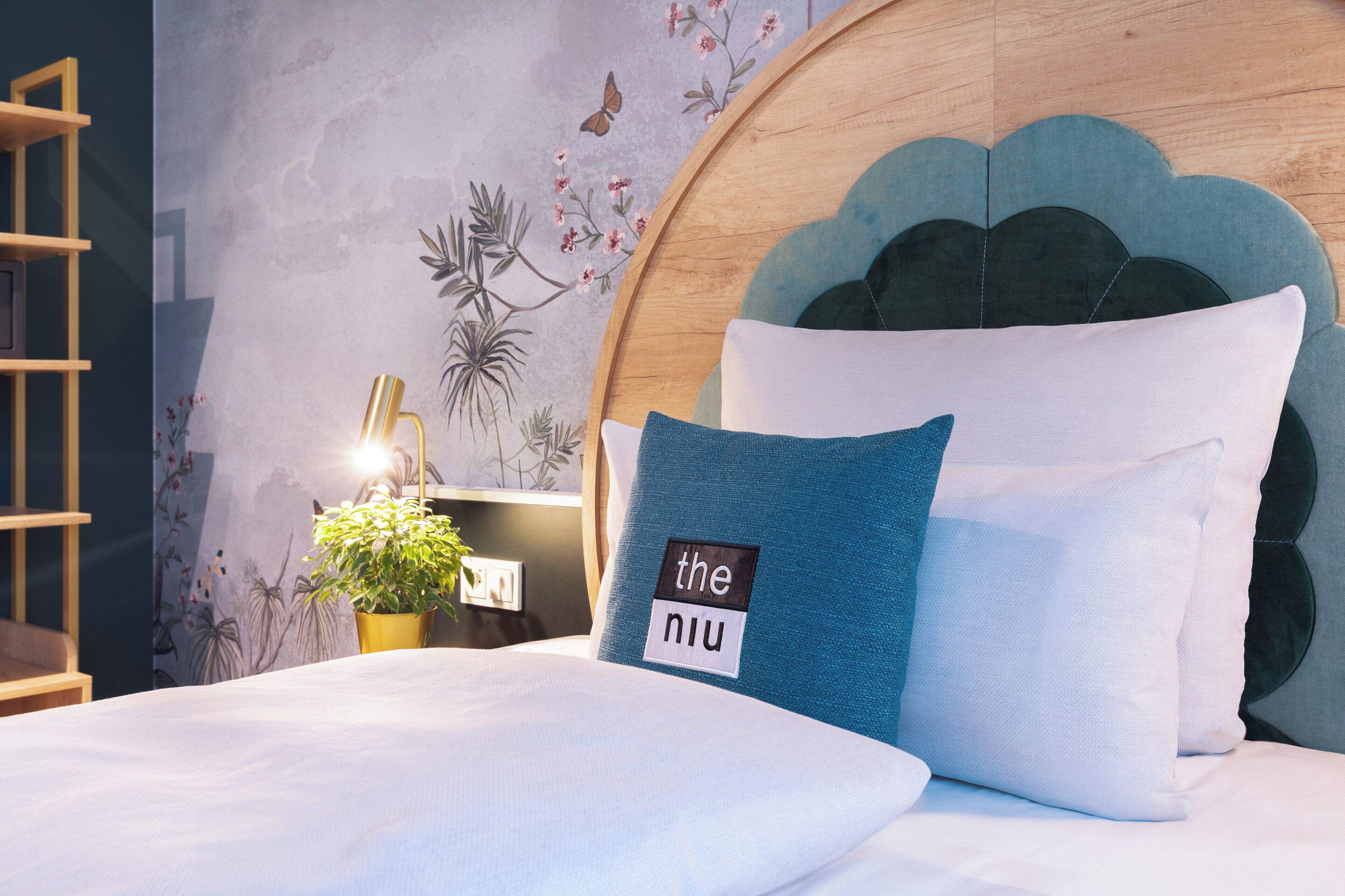 A neatly made bed with a blue pillow and a white pillow, a wooden headboard, a wall with floral and butterfly wallpaper, and a small potted plant on a nightstand.