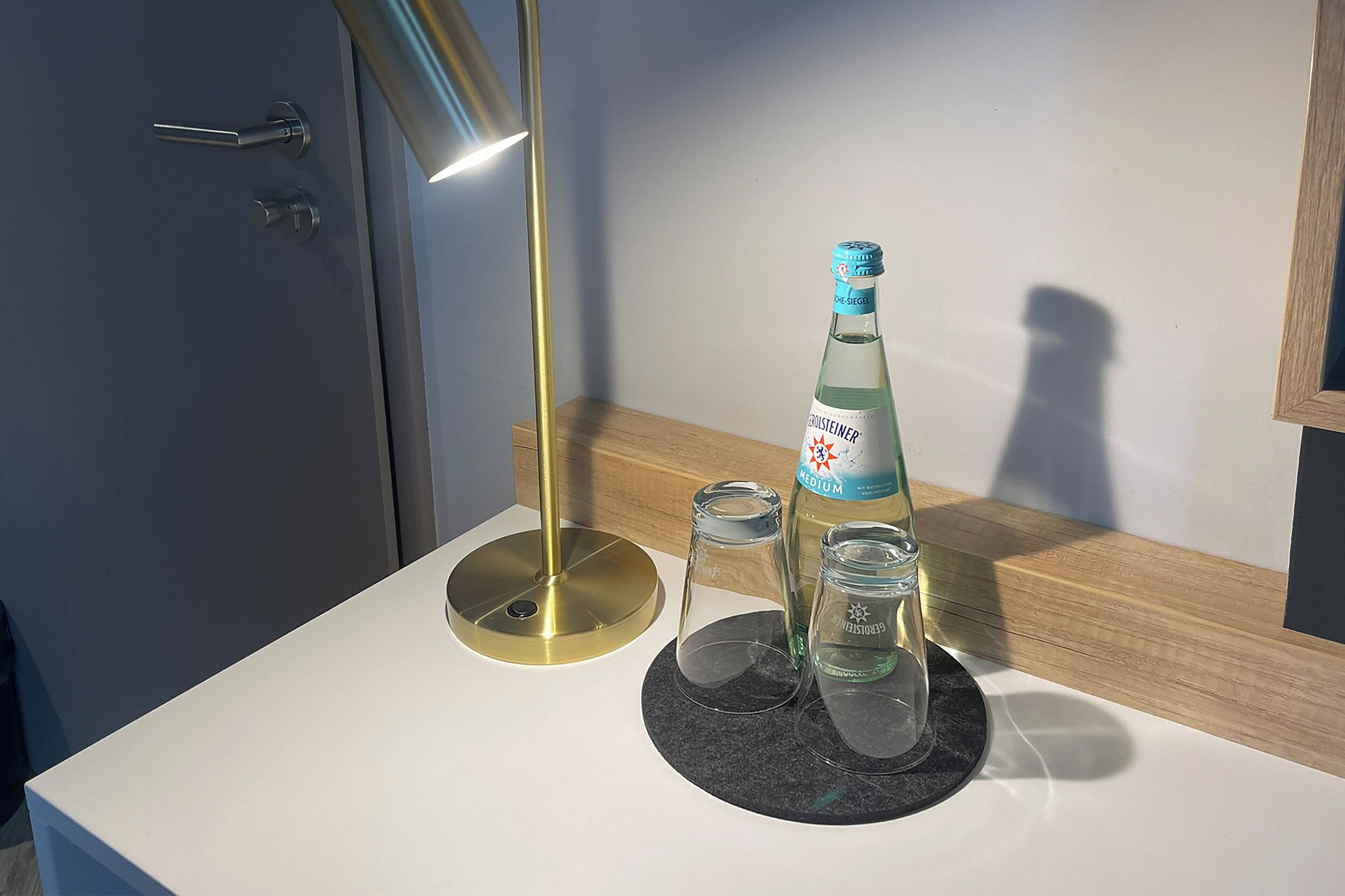 A modern bathroom with a white countertop, a bottle of Groseiner Medium, two clear glasses, and a gold-colored lamp.
